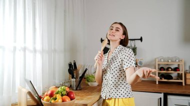 Enerjik beyaz anne kahvaltı için salata pişirirken ve yemek pişirirken elinde kepçeyle şarkı söylüyor. Sağlıklı yemek yapmaya hazırlanırken müziğe yönelen mutlu ev sahibi. Pedagoji.