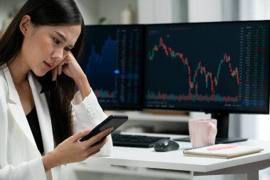 Stresli, Asyalı genç ve güzel iş kadını akıllı telefona odaklanıyor, modern ofisteki bilgisayar ekranında dinamik borsa grafiğinde kar veya zarar etme riskini analiz ediyor. Taktik.