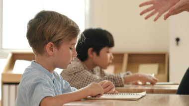 Beyaz tenli ilkokul öğrencisinin tahtada otururken bir nota bakması ve soruya cevap vermesi. Çocuklar derse katılırken genç öğretmen sınıf çalışmasını ya da sınavı açıklar. Pedagoji.