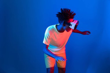 Genç Afrikalı Amerikalı basketbol oyuncusu, metaevren dünyasında dijital fütüristik teknolojiyi birbirine bağlayan mavi neon ışıkta zıplayan VR pozu takıyor. Entrika.