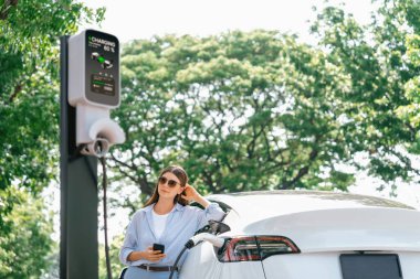 Ulusal Park veya Yaz Ormanı 'ndaki tatil gezisi sırasında EV şarj istasyonundan elektrik aküsü almak için akıllı telefon internet bankacılık uygulaması kullanan genç bir kadın. Yükselt