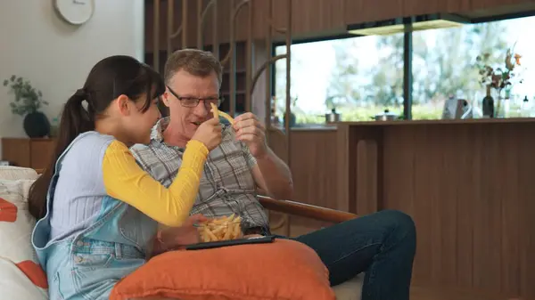 Stock image Grandfather and granddaughter eat french fries as food watch entertainment on TV. Old senior use technology communicate with young generation cross generation gap strengthen family bond. Divergence.