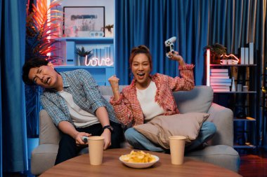 Mutlu çiftin oturduğu kanepe joysticklerle video oyunu oynuyor, meydan okuyan gülümseyen kadın, evdeki modern neon ışıklı oturma odasındaki mücadeleyi kazanmak için yumruk kaldırıyor. İnfobahn.