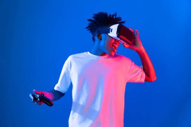 VR kullanan Amerikalı Afrikalı oyun oyuncusu joystick 'i kontrol eden hologram izole edilmiş mavi neon ışıklı duvar ekranı dijital fütüristik teknoloji sanal gerçeklik dünyasına bağlanıyor. Entrika.
