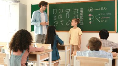 Profesyonel öğretmen konuşuyor ve sınıfta Asyalı çocuğa fikrini açıklıyor. Sınıfın önünde karatahta matematik dersiyle dikilmiş beyaz öğrenci dinleme eğitmeni. Pedagoji.
