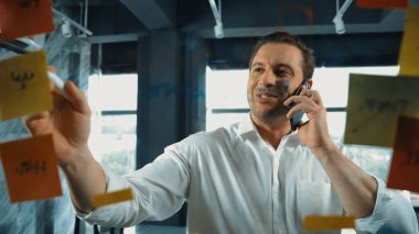 Profesyonel işadamı renkli yapışkan notlara yazılmış pazarlama stratejisi fikrine bakarken müşterilerine telefon ediyor. Proje yöneticisi duvardaki telefon analiz verileriyle konuşuyor. Yönlendirici.