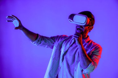 Sanal programı keşfetmek ve girmek için VR gözlük takan heyecanlı bir adam. Yetenekli oyuncular metaevrene dokunurken gündelik giysiler giyer ve neon ışıklı arka planda dururlar. Yaşam tarzı. Sapma.