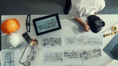 Tablet, dizüstü bilgisayar, güvenlik kaskı ve ekipmanlarla toplantı masasında otururken ev tasarımı için malzeme seçen yetenekli mühendis görüşü. Akıllı mimar proje planına bakıyor. Hizalama.