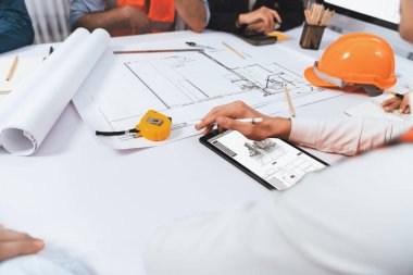 Mimari proje üzerinde birlikte çalışan çeşitli inşaat mühendisi ve müvekkil grupları, inşaat planını gözden geçirip toplantı masasında plan oluşturuyorlar. Tedbirli