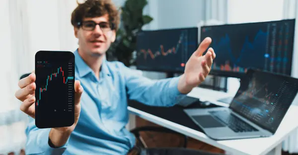 stock image Successful young business trader showing dynamic stock market graph on smartphone, presenting infomation database of currency exchange rate in real time against on pc blurred background. Gusher.