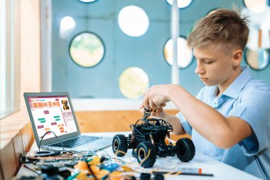 Sarı saçlı çocuk BTMM sınıfında teknoloji öğrenirken kerpeten kullanarak robot araba tamir ediyor. Masaya dizüstü bilgisayar, kumanda, elektrik kablosu, şarj aleti ve robot araç koyun. Öğretim.