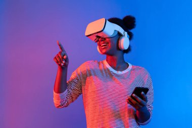 VR kulaklıklı ve akıllı telefonlu genç Afrikalı kadın canlı neon ışıklandırma arka planı sanal gerçekliği ve mobil bağlantı öğrenme teknolojisiyle metaevrene doğru yol alıyor. Entrika.