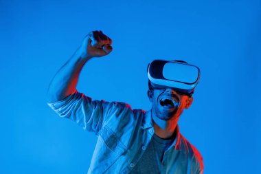 Sanal dünyada spor müsabakalarını izleyen mutlu adam neon ışıklı arka planda VR kulaklık takıyor. Yakışıklı genç, gelecekteki yenilik teknolojisine gözlük kullanarak giriyor. Sapma.