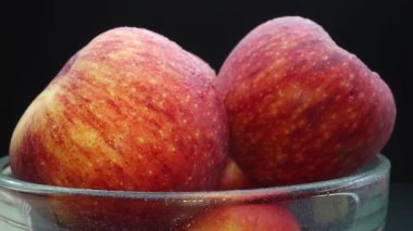 Yakın plan vitrinlerde cam bir kaseye yerleştirilmiş taze kırmızı bir elma görülüyor. Siyah arkaplan, elmaların kusursuz ve pürüzsüz cildini vurgulayan dramatik bir sahne yaratıyor. Geliştirilebilir.