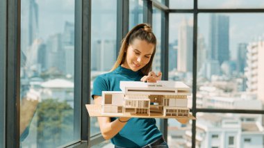 Genç akıllı mimar mühendisi ev modelini incelerken mimari modeli elinde tutuyor. Şehir manzaralı pencerenin yanında dururken profesyonel iç mimar ev inşaatını kontrol ediyor. İzleme