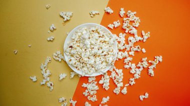 Patlamış mısır, turuncu ve sarı arka planda bir kaseden uçuyor. Beyaz kaseye düşen ve düşen patlamış mısırın yukarıdan görünüşü. Sıradan atıştırmalık konsepti. Poster için tasarım, duvar kağıdı. Pabulum.