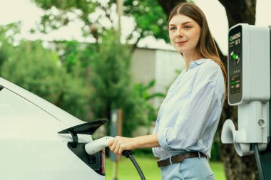 Açık yeşil şehir parkı manzaralı EV şarj istasyonundan genç kadın EV elektrikli araçları şarj ediyor. Çevre dostu şehir ulaşımı ve çevre dostu EV araba seyahati. Yükselt