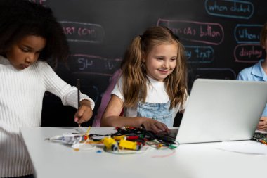 Beyaz önlüklü akıllı kız BTMM sınıfında dizüstü bilgisayar kullanarak robotik teknolojiyi öğreniyor. Mavi tişörtlü öğrenci anakart öğretirken zeki öğrenciler kod okumaya çalışıyor. Etkinlik.