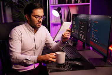 Akıllı bilişim geliştiricisi, bilgisayar ekranındaki yazılım geliştirme kodlaması üzerine kahveyle birlikte çalışıyor, en yeni uygulama projesi programı ile birlikte neon ışıklı siber ofiste çevrimiçi web sitesi verilerini güncelliyor. Tahmin et..