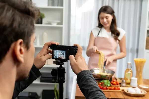 stock image Cameraman recording to woman in chef influencer host cooking spaghetti with meat topped tomato sauce surrounded ingredients recipe, presenting special dish healthy food at modern studio. Postulate.