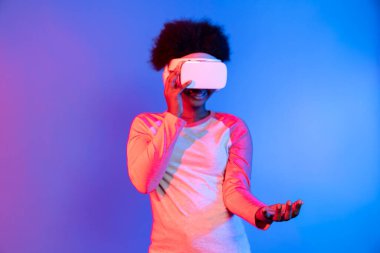 Genç Afrikalı, metaevren üzerinden VR gözlük takıyor ve elinde mavi pembe neon ışık grafikli dijital yenilik üzerine veri öğrenme uygulaması tutuyor. Entrika.