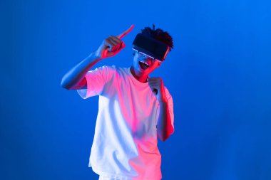 Genç Afrikalı Amerikalı oyuncu sanal gerçeklik dünyasını dijital fütüristik teknolojiye bağlayan mavi neon ışıklandırma duvarında sanal dünyada VR kullanarak bir hologramda yumruk kaldırıyor. Entrika.