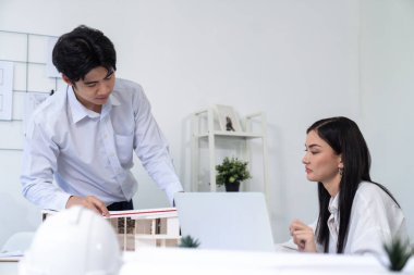 Profesyonel Asyalı erkek mimar ev modelinin uzunluğunu ölçmek için cetvel kullanırken, beyaz tenli genç meslektaşı ev modeliyle toplantı masasındaki verileri analiz etmek için dizüstü bilgisayar kullanıyor. Tertemiz..