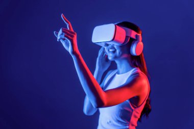 Siberpunk neon ışıklı cihazlarla çevrili zeki bir kadın metaevrene bağlı VR kulaklık takıyor, gelecekçi siber uzay teknolojisi. Sanal objeyi işaret eden bir kadın. Halüsinasyon.