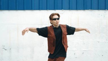 Beyaz arka planda break dans pratiği yapan yakışıklı bir sokak dansçısı. Hip hop tarzı giyinen sporcu, mavi arka planlı bir sokakta müzik eşliğinde ayak sesleri çıkarıyor. Açık hava sporu 2024 hiphop..
