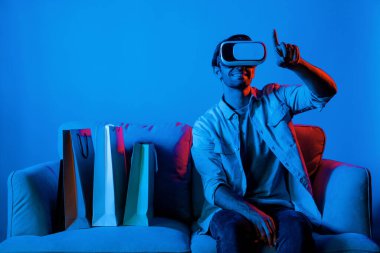 Video kameralı beyaz bir adam kanepeye oturmuş alışveriş torbalarıyla işaret ediyor. VR teknolojisi ve evde sanal gözlük kullanarak online alışveriş yapan kişi. Sapma.