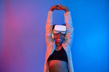 VR giyen genç Afrikalı spor kadın, dijital fütüristik teknolojiyi sanal gerçekliğe bağlayan parlak mavi neon ışıkları üzerinde poz veriyor. Entrika.