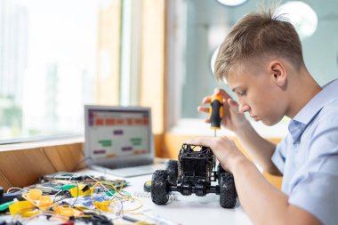 Sarı saçlı okul çocuğu, BTMM sınıfında robotik araç tamir eden bir tornavida kullanıyor. Masaya dizüstü bilgisayar, kumanda, elektrik kablosu, şarj aleti ve robot araç koyun. Öğretim.