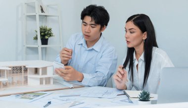 Profesyonel Asyalı erkek mimar ev modelinin uzunluğunu ölçmek için cetvel kullanırken, beyaz tenli genç meslektaşı ev modeliyle toplantı masasındaki verileri analiz etmek için dizüstü bilgisayar kullanıyor. Tertemiz..