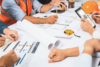 Mimari proje üzerinde birlikte çalışan çeşitli inşaat mühendisi ve müvekkil grupları, inşaat planını gözden geçirip toplantı masasında plan oluşturuyorlar. Tedbirli