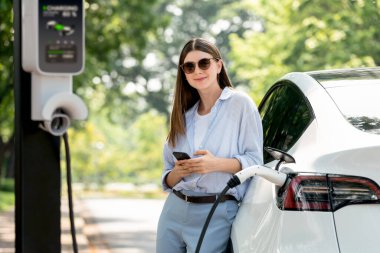 Ulusal Park veya Yaz Ormanı 'ndaki tatil gezisi sırasında EV şarj istasyonundan elektrik aküsü almak için akıllı telefon internet bankacılık uygulaması kullanan genç bir kadın. Yükselt