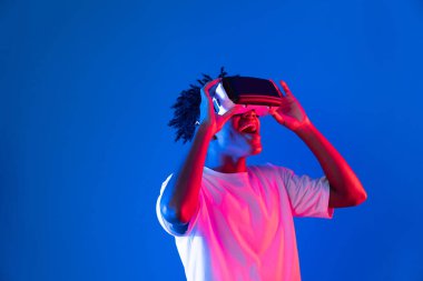 Sanal gerçekliği dijital fütüristik teknolojiye bağlayan pembe mavi neon duvarda VR 'dan bakan genç Afrikalı Amerikalı' ya şaşırdım. Entrika.