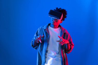 VR kullanan genç Afro-Amerikalı, boş uzay ürünlerini işaret ederek metaevren dünyasındaki mavi, neon ışıklı duvar fütüristik teknolojisi hakkında detaylı bilgi veriyor. Entrika.