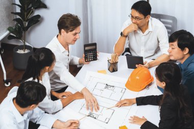 Mimari proje üzerinde birlikte çalışan çeşitli inşaat mühendisi ve müvekkil grupları, inşaat planını gözden geçirip toplantı masasında plan oluşturuyorlar. Tedbirli