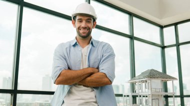 Sıradan kıyafetli mimar mühendisinin portresi kollarını kavuştururken kameraya gülümsüyor. Mühendis kameraya bakıyor ve güvenlik kaskı takarken ev modelinin yanında kollarını kavuşturmuş oturuyor. İzleme
