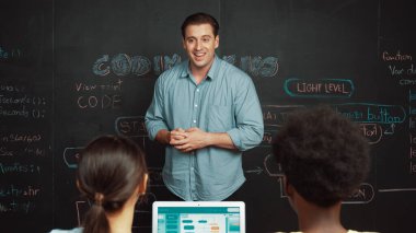 Tahtada çeşitli öğrencilere mühendislik kodları öğreten yakışıklı bir öğretmen. Mutlu çok kültürlü liseli çocuklar dizüstü bilgisayar kullanırken öğreniyor ya da programlama veya kodlama sistemini öğreniyor. Öğretim.