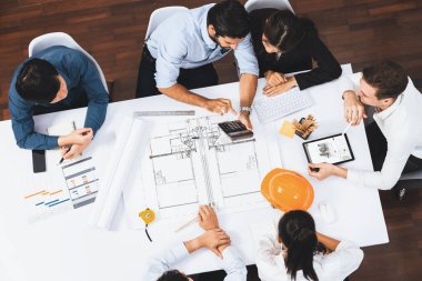 Mimari proje üzerinde birlikte çalışan, inşaat planını gözden geçiren ve toplantı masasında plan oluşturan çeşitli inşaat mühendisi ve müşteri gruplarından oluşan üst düzey bir pankart. Tedbirli