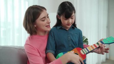 Kafkasyalı bir anne evde akustik müzik öğretirken ukulele çalan mutlu kız. Sevimli çocuk enstrüman öğreniyor. Kafkasyalı mutlu anne ve kız birlikte vakit geçiriyorlar. Pedagoji.