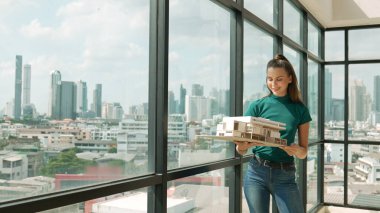 Genç akıllı mimar mühendisi ev modelini incelerken mimari modeli elinde tutuyor. Şehir manzaralı pencerenin yanında dururken profesyonel iç mimar ev inşaatını kontrol ediyor. İzleme
