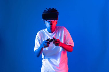 VR kullanan Amerikalı Afrikalı oyun oyuncusu joystick 'i kontrol eden hologram izole edilmiş mavi neon ışıklı duvar ekranı dijital fütüristik teknoloji sanal gerçeklik dünyasına bağlanıyor. Entrika.