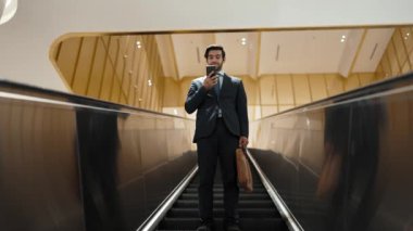 Profesyonel bir iş adamı yürüyen merdiven kullanırken telefonla konuşuyor. Beyaz tenli yönetici, pazarlama fikri ya da planı rapor etmek ve tartışmak için iş ekibiyle konuşuyor. Ön manzara. Sevinçli..