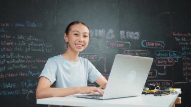Akademik liseli kız BTMM teknoloji sınıfında kameraya bakarken dizüstü bilgisayar kullanarak kod yazıp çalışıyor. Öğrenci gülümsemesi masaya yerleştirilmiş elektronik aletlerle kameraya. Öğretim