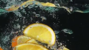 Arkaplanlı cam bir kase suda yüzen portakal dilimleri. Portakal dilimlenmiş makrografi. Ayrı bir karanlık zemin ile berrak suya düşüyor. Kapatın. Ağır çekim. Pabulum.