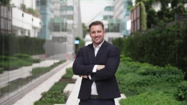 Gülümseyen beyaz iş adamı kollarını kavuşturup şehir merkezindeki kameraya bakıyor. Yönetici, yeşil şehirde takım elbise giyerken kolları bağlı bir şekilde dikiliyor. Yeşil çevre. Urbane.