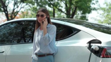 Doğal orman ya da milli parktaki araba yolculuğu sırasında elektrikli araba pili şarj ederken kadın telefonda konuşuyor. Tatil ve tatil boyunca çevre dostu seyahat. Yükselt