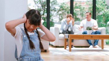 Stresli ve mutsuz genç kız köşeye kıvrılıp kulaklarını kapatarak arka planda ebeveyninin tartışmasını engelliyor. Evde aile içi şiddet ve travmatik çocukluk depresyona dönüşür. Eşzamanlama